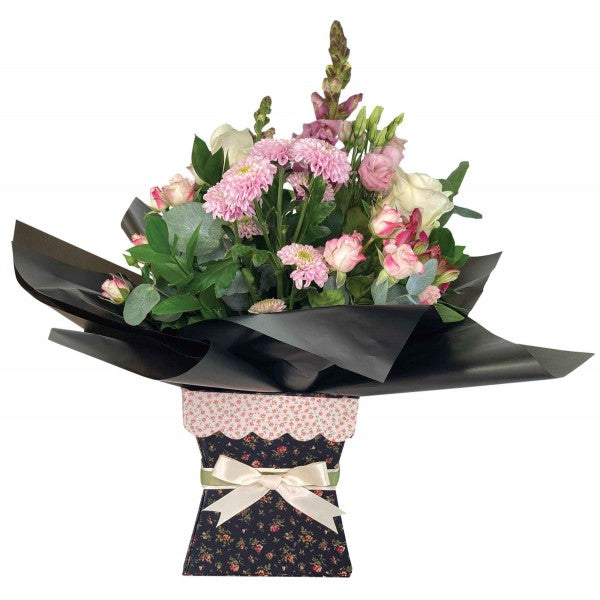 A floral bouquet with pink and white flowers, green foliage, and black wrapping paper, presented in a decorative dark floral box with a cream ribbon bow on a white background.