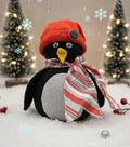 Decorative penguin with a red hat and striped scarf in a snowy scene with Christmas trees.